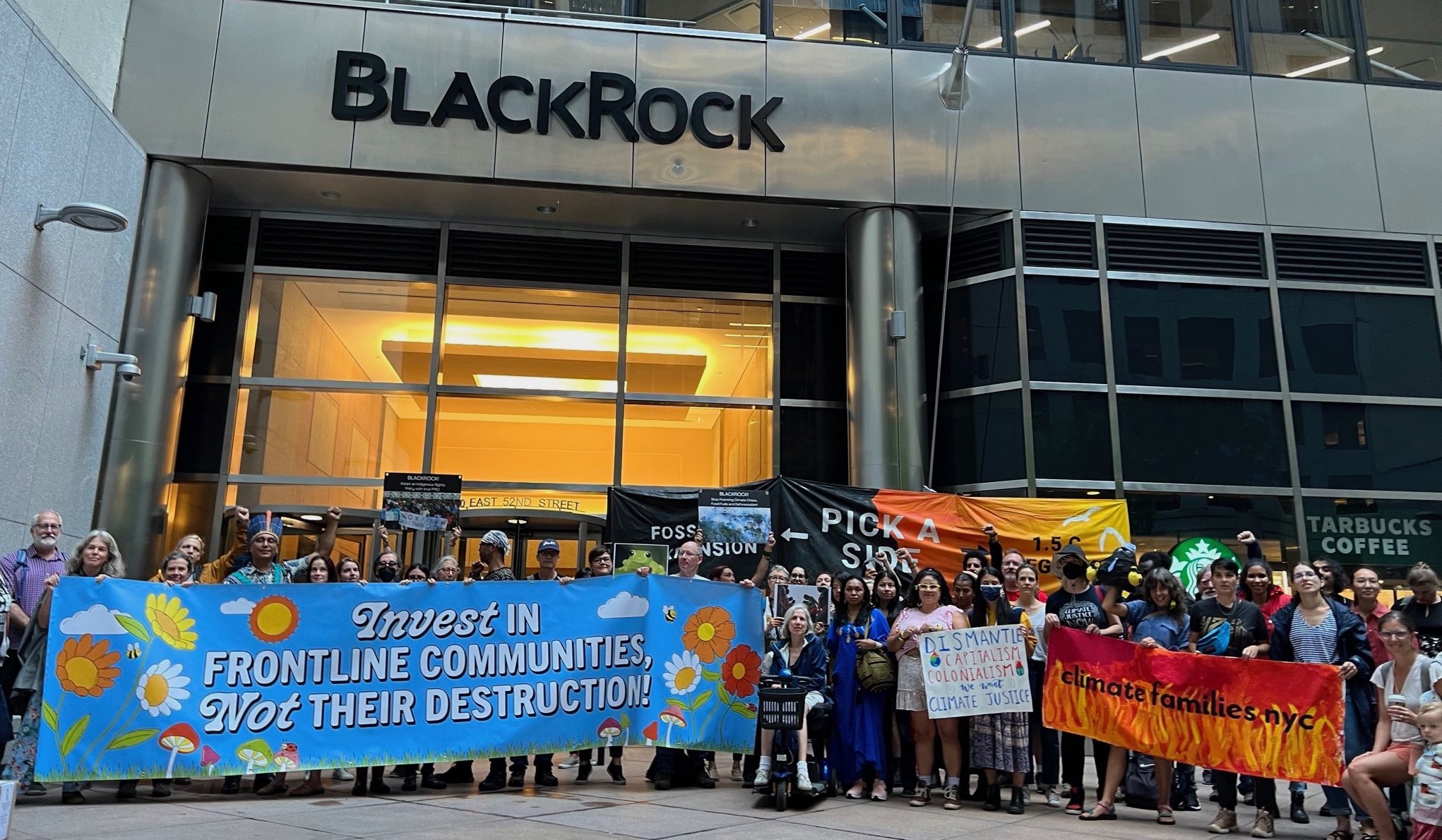 A large group of protestors holding signs and banners gather in front of BlackRock's NYC headquarters.