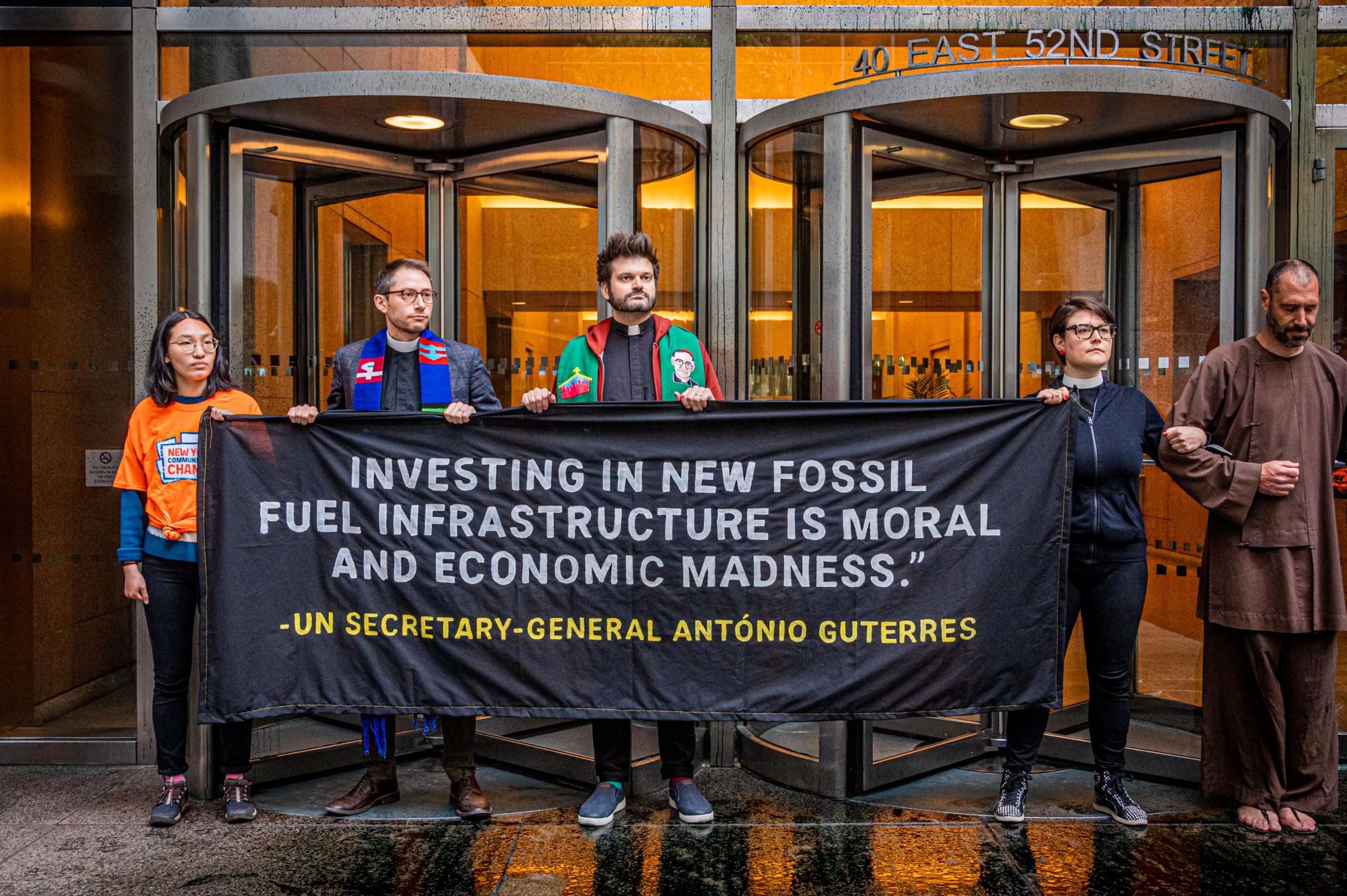 6 activists block the entrance of BlackRock's NYC office holding signs with faces of BlackRock leadership on them that all read "pick a side." They are also holding a large black banner that has a quote from U.N. Secretary-General Guterres. It reads "Investing in new fossil fuel infrastructure is moral and economic madness." Photo by Erik McGregor.