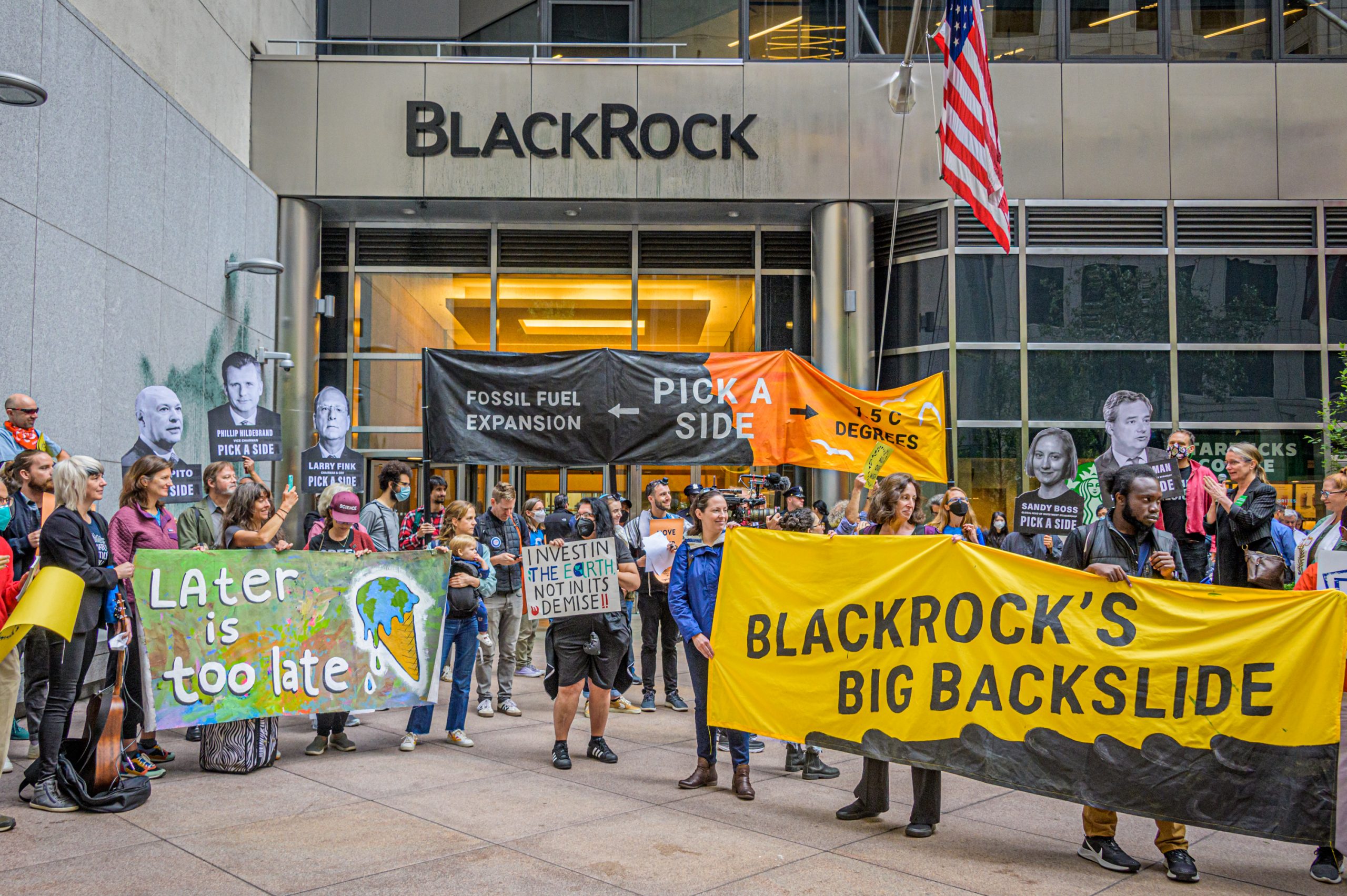 A crowd of activists with several large banners and signs stand in front of BlackRock's headquarters. The signs read "Fossil Fuel expansion or 1.5C degrees, pick a side", "Later is too late", "BlackRock's Big Backslide", and "New York Communities for Change." Photo by Erik McGregor.